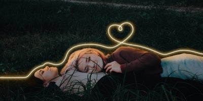 Två tjejer ligger i gräset och en glödande tråd formar ett hjärta över dem