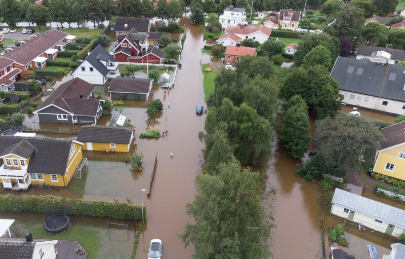 Flygbild av översvämmat bostadsområde. Hus, vägar och trädgårdar står under vatten, människor vadar på gatan.